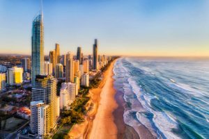The golden morning sun rises over the Pacific Ocean, casting a warm orange and pink glow onto the sleek skyscrapers of Surfers Paradise. The Q1 building stands tall in the center, its glass facade reflecting the sunrise, while gentle waves lap onto the wide, empty beach in the foreground