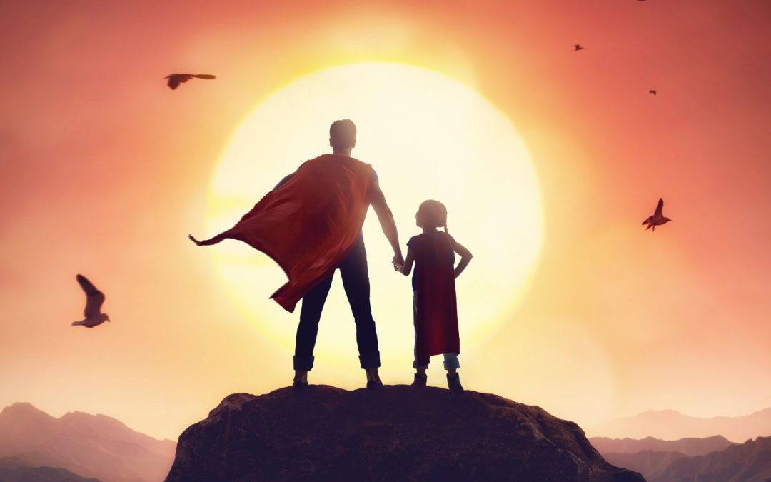 Father with a cape holding daughter's hand in top of the mountain, standing against the sun